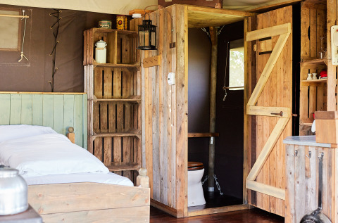 Interno rustico di una tenda safari con letto in legno, scaffali, bagno privato e luce naturale.
