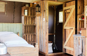 Intérieur rustique de tente safari avec lit en bois, étagères, salle de bain privée et lumière naturelle.