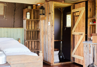 Interno rustico di una tenda safari con letto in legno, scaffali, bagno privato e luce naturale.
