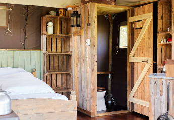 Interno rustico di una tenda safari con letto in legno, scaffali, bagno privato e luce naturale.