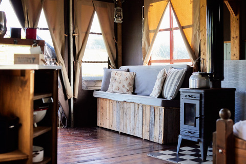Interior view of a safari tent lounge with wooden bench seating, pillows, wood stove, and sunny windows.