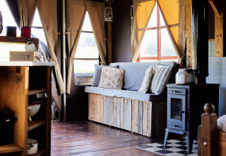 Interior view of a safari tent lounge with wooden bench seating, pillows, wood stove, and sunny windows.