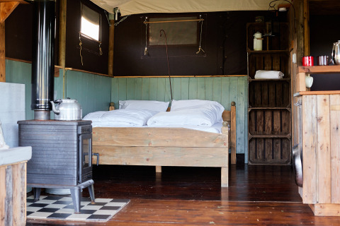 Intérieur d'une tente safari avec lit double en bois, poêle, étagères rustiques et sol en bois.