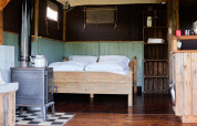 Intérieur d'une tente safari avec lit double en bois, poêle, étagères rustiques et sol en bois.