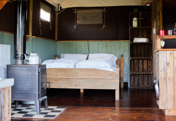 Interior of a safari tent featuring a wooden double bed, stove, rustic shelves, and wooden flooring.