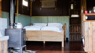 Het interieur van een safaritent met houten tweepersoonsbed, houtkachel, planken en houten vloer.