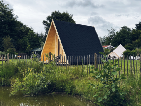 A-Frame damhus i træ med sort tag, indhegnet og beliggende ved en dam, omgivet af grønt og træer.