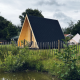 A-Frame damhus i træ med sort tag, indhegnet og beliggende ved en dam, omgivet af grønt og træer.