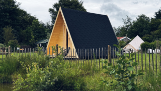Cabane A-Frame pond house en bois, toit noir, clôture, près d'un étang entouré de verdure et d’arbres.