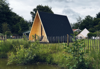 Cabane A-Frame pond house en bois, toit noir, clôture, près d'un étang entouré de verdure et d’arbres.