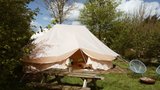 Una tenda safari Bell Tent Emperor tra alberi verdi con tavolo da picnic e sedie da esterno davanti.