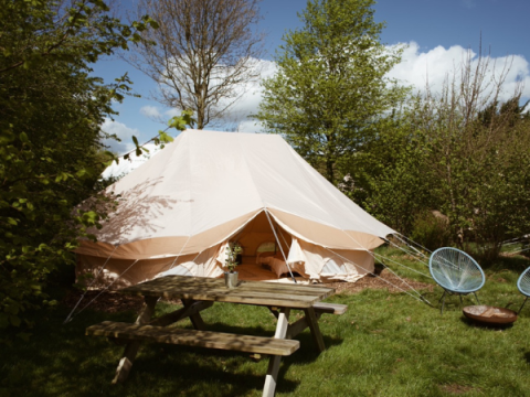Une tente safari Bell Tent Emperor installée parmi des arbres verts, avec table de pique-nique et chaises.