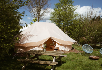 En Bell Tent Emperor-safari telt omgivet af grønne træer med et picnicbord og udendørs stole foran.
