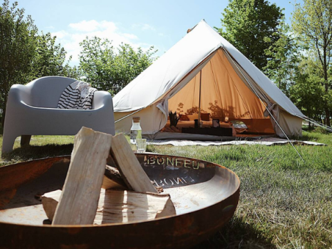 Billede af et Bell Tent 500 safari-telt med åbning, lejrbål og stol i en grøn, solrig natur.