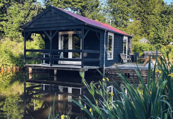 Zwart houten huisje aan een vijver tussen het groen met terras, stoelen en tafel buiten.