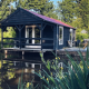 Sort træhytte ved en dam omgivet af grøn natur, campingterrasse med stole og bord i det fri.