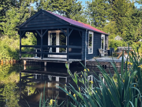 Schwarze Holzhütte am Teich, umgeben von grüner Natur, Terrasse mit Stühlen und Tisch im Freien.
