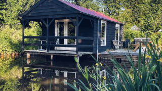 Black wooden cabin by a pond, surrounded by lush greenery, outdoor deck with chairs and a table.
