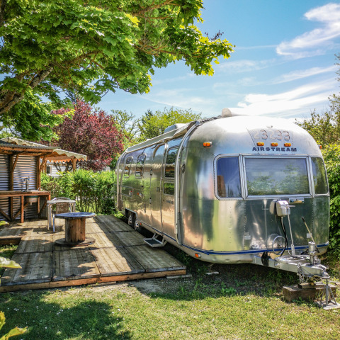 Vintage Airstream-campingvogn i solen med træterrasse og have ved Camping Le Château, Auvergne-Rhône-Alpes.