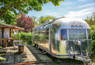 Vintage Airstream-campingvogn i solen med træterrasse og have ved Camping Le Château, Auvergne-Rhône-Alpes.