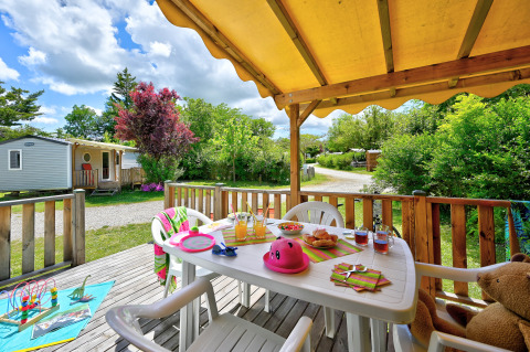 Terrasse mit Esstisch, Stühlen und Kinderspielzeug inmitten grüner Natur im Camping Le Château, Frankreich.
