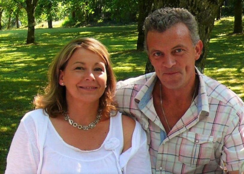 A smiling couple sitting outdoors in the sun at Camping Le Château, a holiday park in Auvergne-Rhône-Alpes, France.