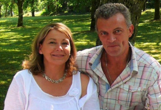 A smiling couple sitting outdoors in the sun at Camping Le Château, a holiday park in Auvergne-Rhône-Alpes, France.