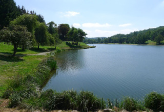 Ruhiger See mit Bäumen und grüner Wiese im Ferienpark Camping Le Château, Auvergne-Rhône-Alpes, Frankreich.
