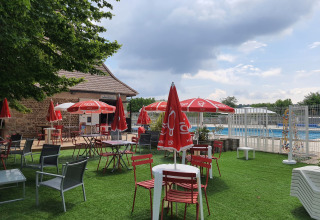 Tavoli e sedie all'aperto con ombrelloni rossi vicino alla piscina al Camping Le Château, Auvergne-Rhône-Alpes.