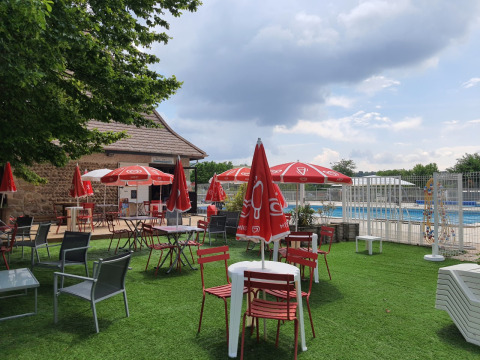 Outdoor tables, chairs, and red umbrellas by the pool at Camping Le Château in Auvergne-Rhône-Alpes, France.