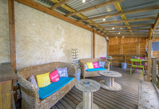 Überdachte Terrasse im Camping Le Château, Auvergne-Rhône-Alpes, mit Loungesofas und bunten Kissen.