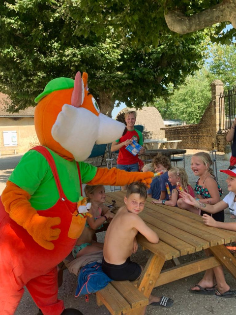 Bambini si divertono con una mascotte colorata a un tavolo da picnic al Camping Le Château, Auvergne-Rhône-Alpes.
