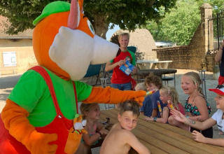Children laughing with a colorful mascot at a picnic table in Camping Le Château, Auvergne-Rhône-Alpes, France.