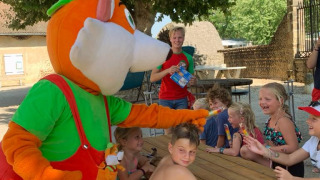Bambini si divertono con una mascotte colorata a un tavolo da picnic al Camping Le Château, Auvergne-Rhône-Alpes.