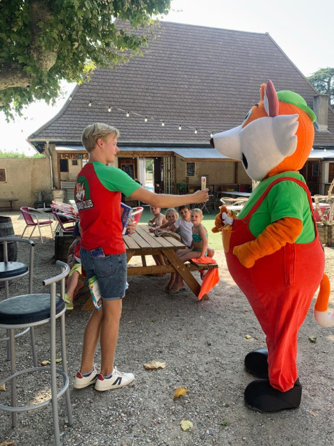 Un joven con camiseta roja saluda a una mascota de zorro cerca de una mesa de picnic al aire libre con niños.