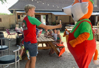 A person in a red shirt meets a fox mascot near a wooden picnic table with kids seated outdoors at a park.