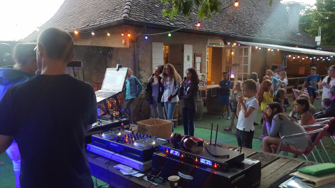 Udendørs aftenarrangement på Camping Le Château med DJ-udstyr, børn og voksne under lyskæder.