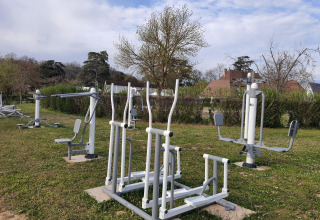 Outdoor fitness equipment set on grass in a holiday park, with trees and houses in the background at Camping Le Château.