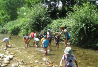 Children playing in a shallow stream near lush greenery at Camping Le Château, Auvergne-Rhône-Alpes, France.