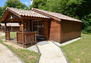 Holzhütte mit überdachter Terrasse im Camping Le Château, umgeben von Bäumen in Auvergne-Rhône-Alpes, Frankreich.