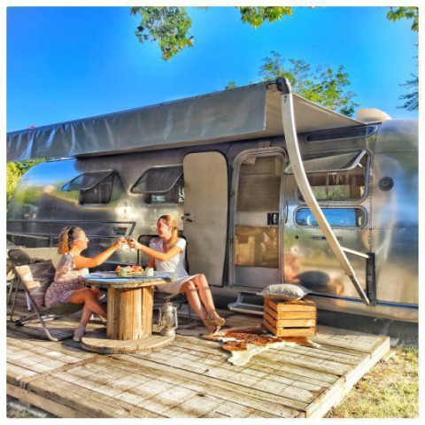 To kvinder nyder mad og drikke udenfor en amerikansk Airstream caravan med privat spa på en solrig dag.