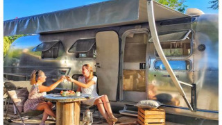 Two women enjoying food and drinks outside an American Airstream caravan with a private spa on a sunny day.