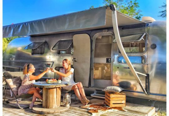 Dos mujeres disfrutan de comida y bebidas frente a una caravana Airstream americana con spa privado al sol.