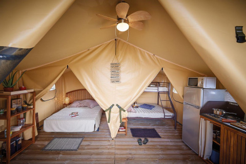 Innenraum eines Safari-Zeltes Lodge Bali Confort auf Camping Le Château, mit Betten, Küche und Regalen.