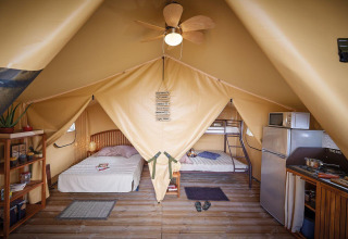 Interior de tienda safari Lodge Bali Confort en Camping Le Château, con camas, cocina y estantería.