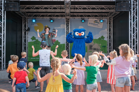 Kinderen dansen voor een artiest en een blauwe mascotte tijdens een show op een glampingterrein.
