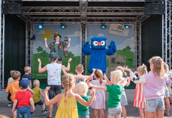 Kinder tanzen vor einer verkleideten Person und einem blauen Maskottchen auf einer Glamping-Bühne.
