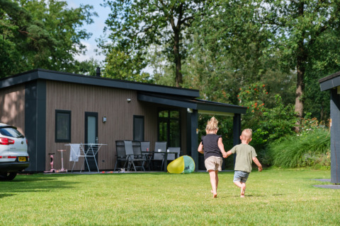 Two children run hand in hand on the grass in front of Forest Lodge DeLuxe at Kampeerdorp de Zandstuve, Netherlands.