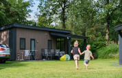 Zwei Kinder laufen Hand in Hand auf dem Rasen vor der Forest Lodge DeLuxe im Kampeerdorp de Zandstuve, Niederlande.