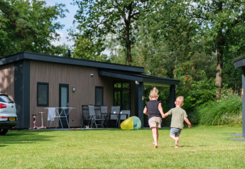 Zwei Kinder laufen Hand in Hand auf dem Rasen vor der Forest Lodge DeLuxe im Kampeerdorp de Zandstuve, Niederlande.
